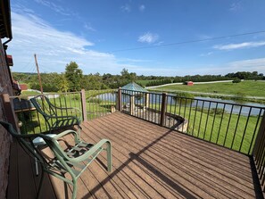 Eine Holzterrasse mit Metallgeländern, zwei grüne Stühle und Blick auf ein grünes Feld sowie ein kleines Gewässer.