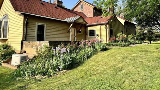 A two-story house with a red roof, yellow siding, and a well-maintained front yard with flowers and a lawn.