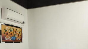A neatly made bed with white bedding, a remote control, and a smartphone on the bed. There is a wall-mounted air conditioner and a framed picture of sunflowers on the wall.