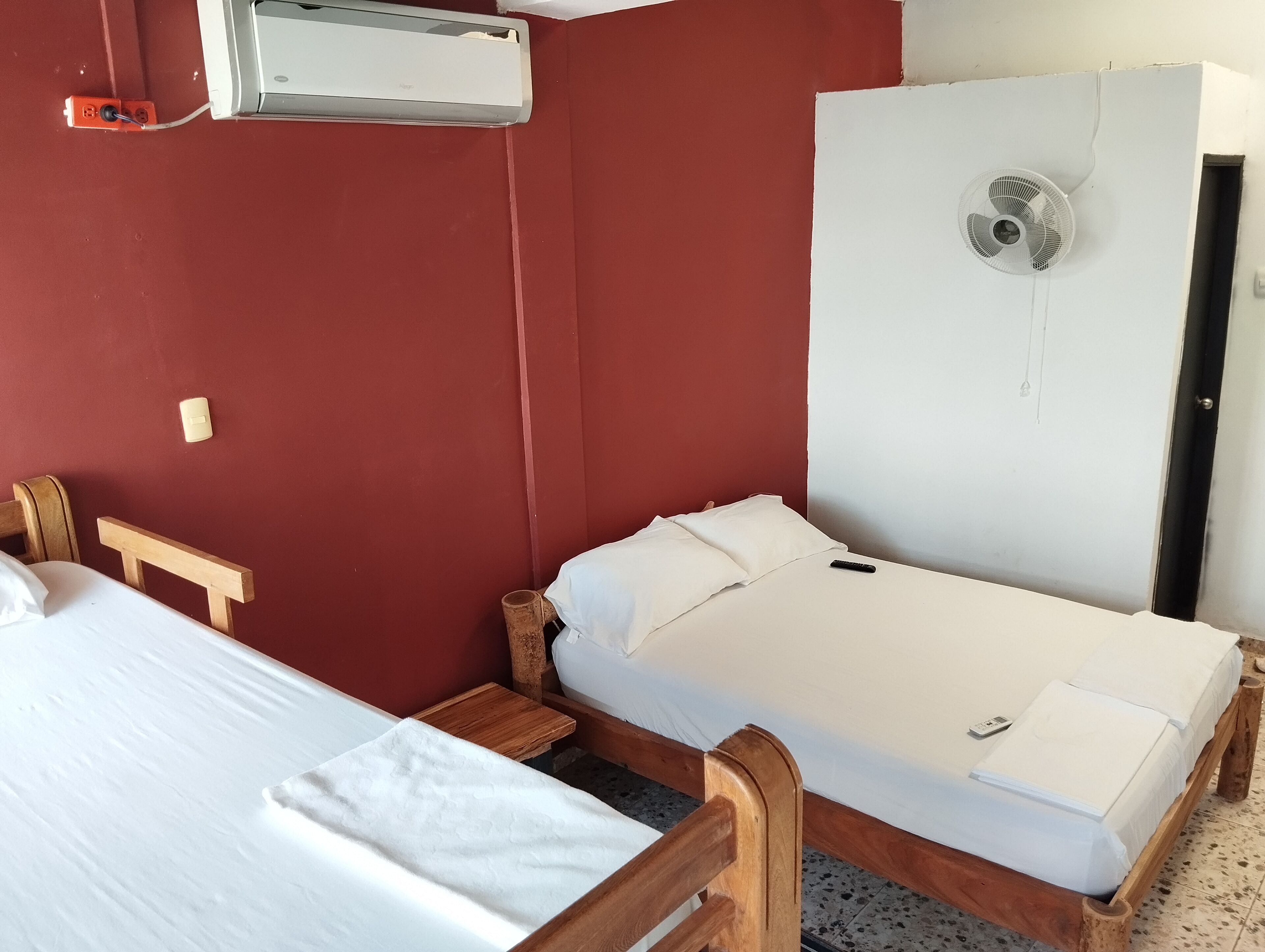 Una habitación con dos camas de madera, una pared roja, un equipo de aire acondicionado y una puerta blanca con un ventilador.