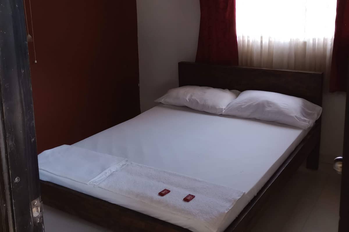 Un lit simple avec une literie et des oreillers blancs, une tête de lit en bois, une fenêtre avec des rideaux rouges et un ventilateur fixé au mur.
