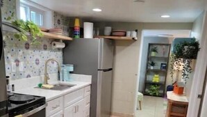 Une cuisine avec des meubles blancs, un réfrigérateur en acier inoxydable, un évier et une cuisinière. Il y a un tapis tissé sur le sol et une étagère avec des plantes.