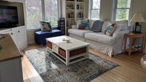 Una sala de estar con un sofá blanco, un reposapiés azul y una mesa de centro. Hay estanterías, un televisor y una ventana con vistas a árboles.