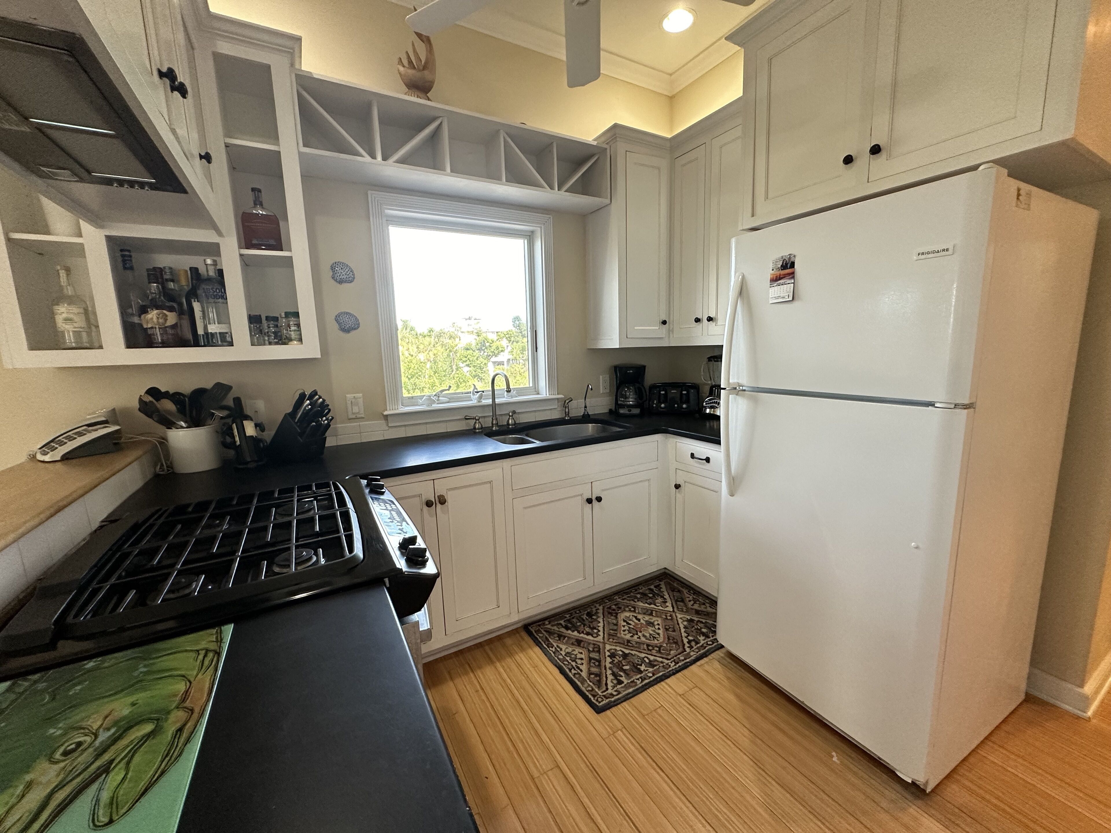 A kitchen with white cabinets, a black countertop, a stove, a refrigerator, and a window with a view of greenery outside.