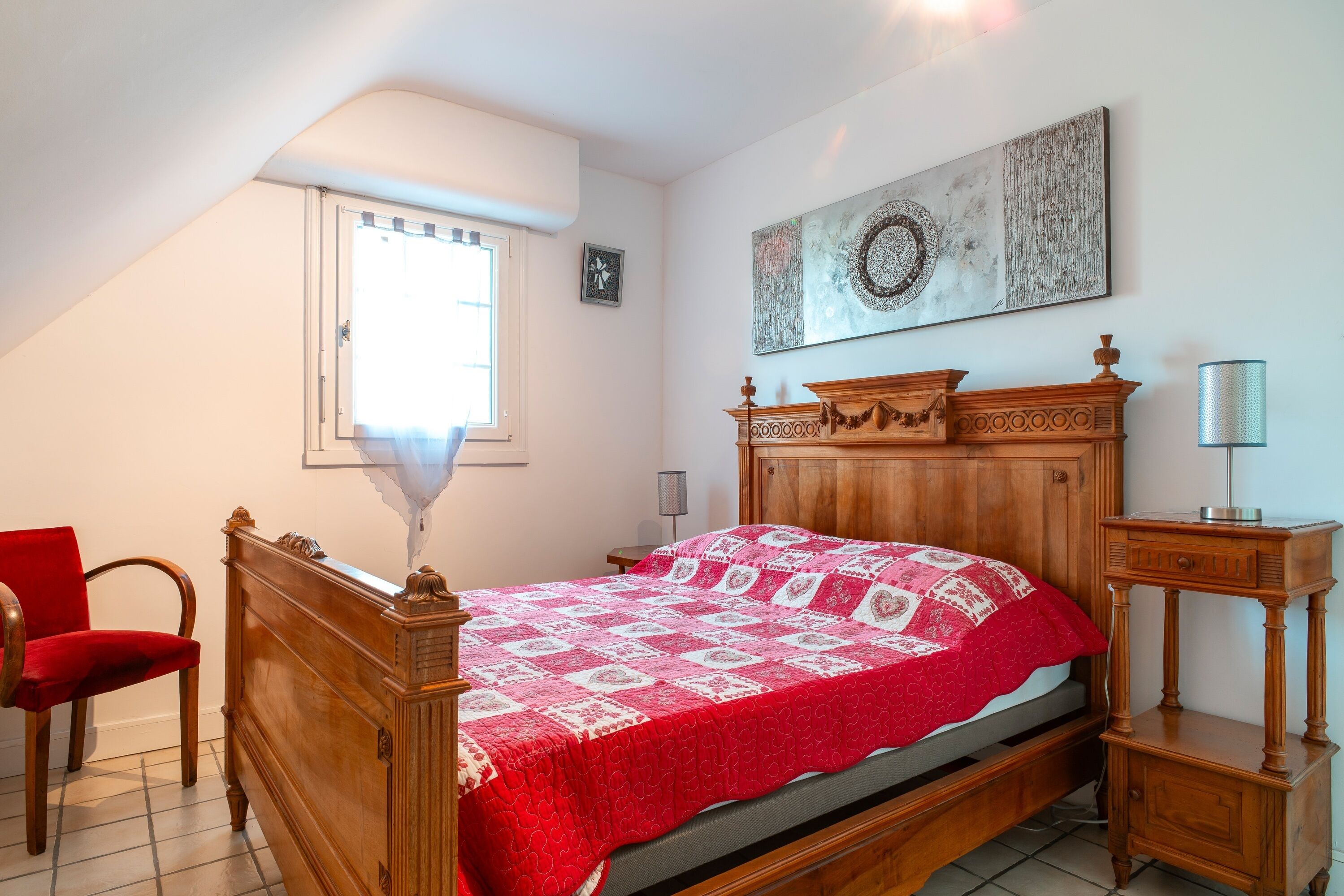 Une chambre à coucher avec un lit en bois, une chaise rouge, une table de chevet et une fenêtre avec des rideaux.