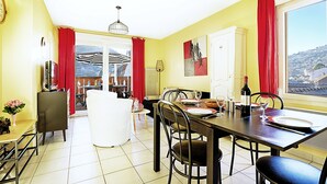 A dining area with a table set for two, chairs, a red curtain, and a view of a cityscape through a large window. - Comme Chez Soi - Vue montagne et garage privé (La Bresse)