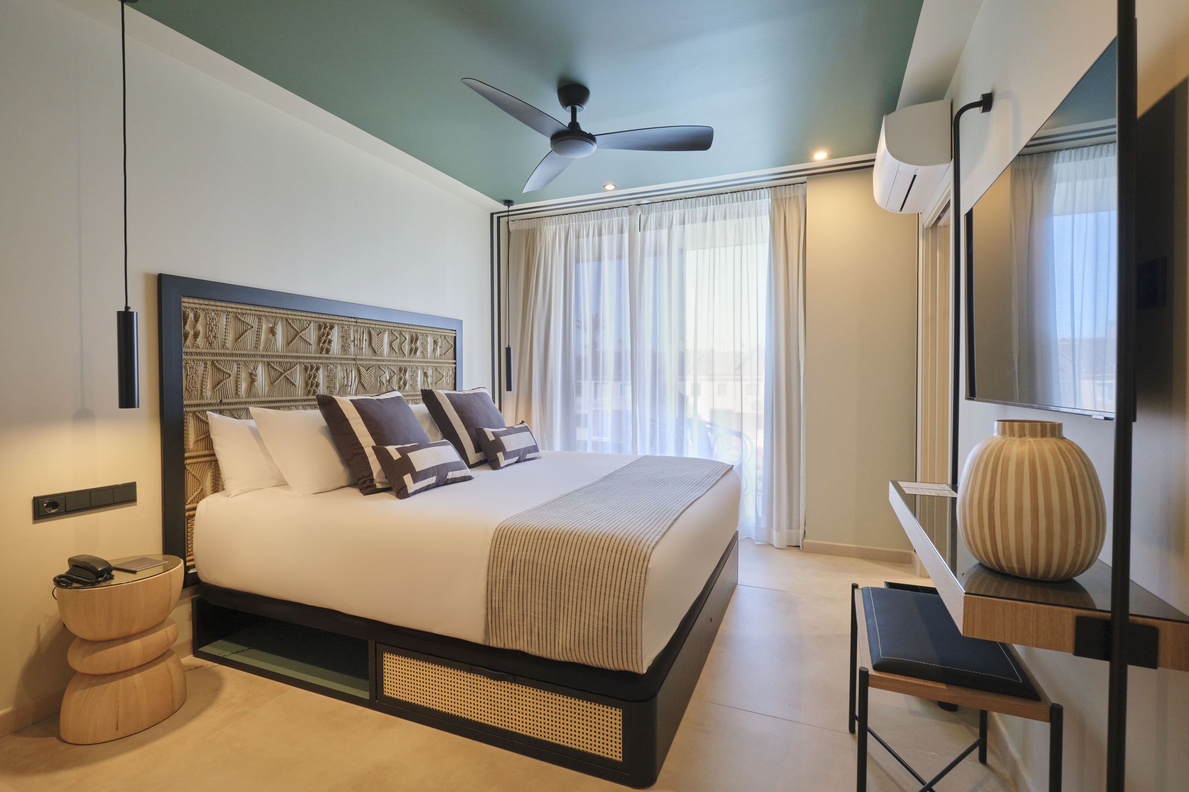 A modern hotel room with a large bed, a ceiling fan, a desk with a vase, and a view of the outside through the window.