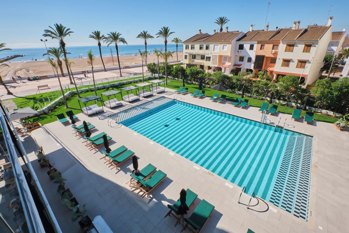 Ein großer Pool im Freien mit Liegestühlen und Sonnenschirmen, umgeben von Palmen und Blick auf Strand und Meer.