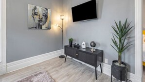 Una sala de estar moderna con una pared gris, un televisor de pantalla plana montado sobre un mueble bajo de madera oscura, una lámpara de pie y un cuadro abstracto grande.
