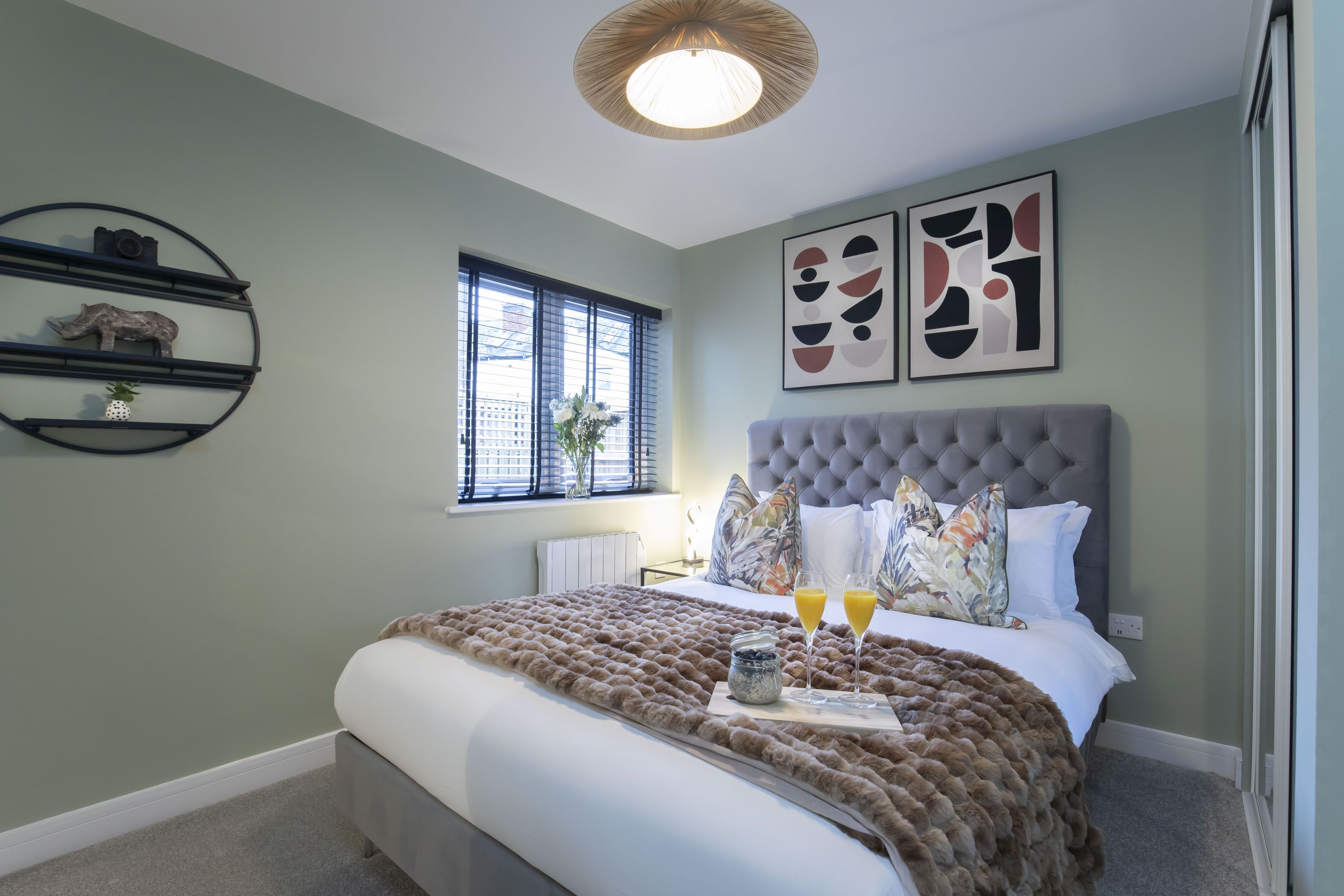 Une chambre à coucher bien aménagée, avec une tête de lit capitonnée, un grand lit recouvert d’une couverture marron texturée, deux tables de chevet ornées d’objets décoratifs et une fenêtre avec des stores.