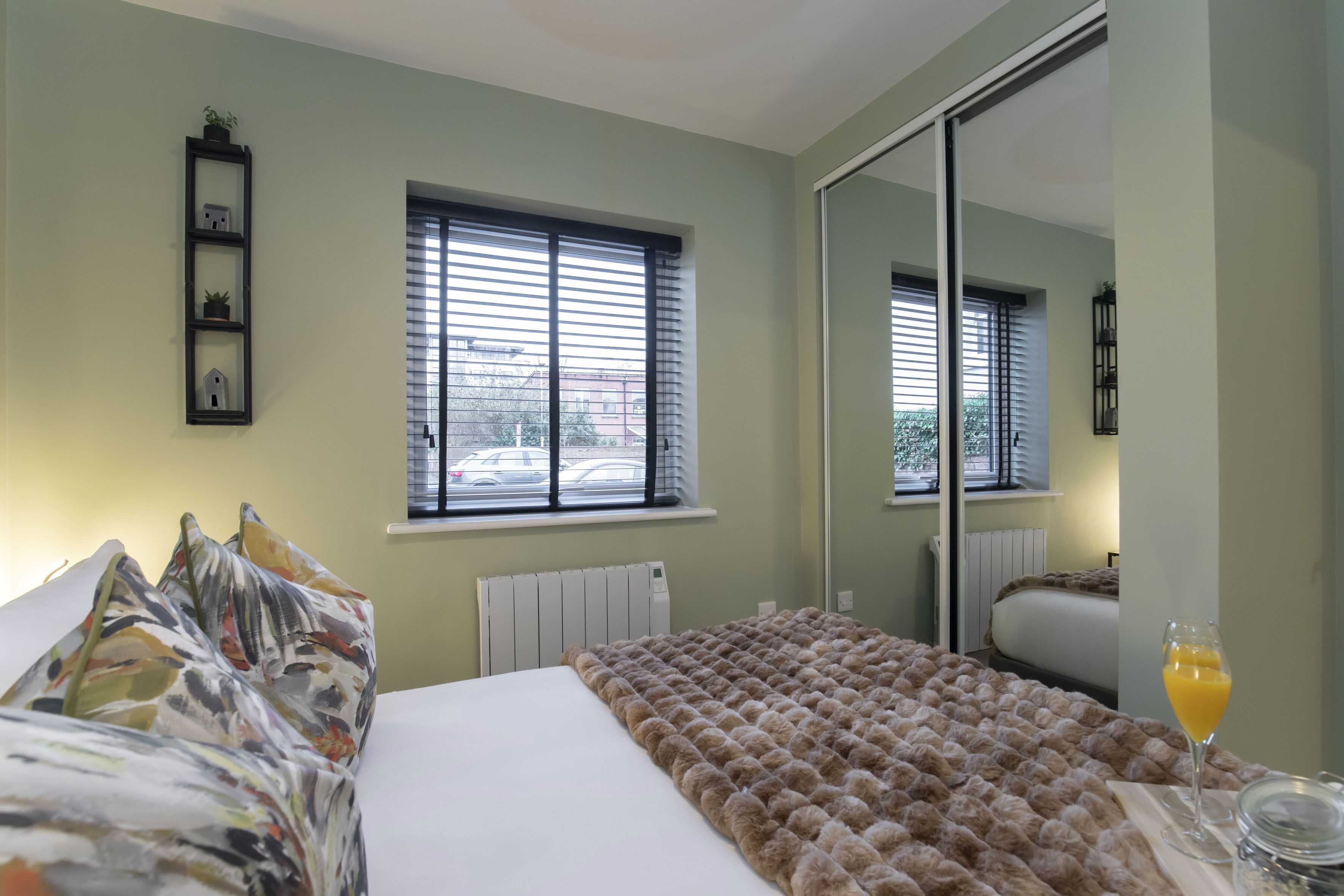 A hotel room with a bed, a window with blinds, a radiator, a mirror, and a glass of orange juice on the nightstand.