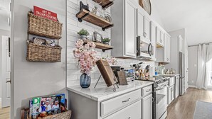 A modern kitchen with white cabinetry, a marble countertop, and various kitchen appliances. There are wicker baskets on the wall and a vase with flowers on the counter. - Edelweiss Chic Retreat /Grand Park/Ruoff/ Extended Stays Accepted
 (Noblesville)
