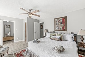 A bedroom with a large bed, a ceiling fan, a door leading to another room, a framed artwork on the wall, and a bedside table with a lamp.
