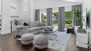 A modern living room with a grey sofa, ottomans, a glass coffee table, and a view of the outdoors.