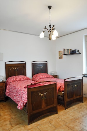 A bedroom with two wooden beds, a chandelier, a bedside table with a lamp, a shelf with books, and a framed picture on the wall.