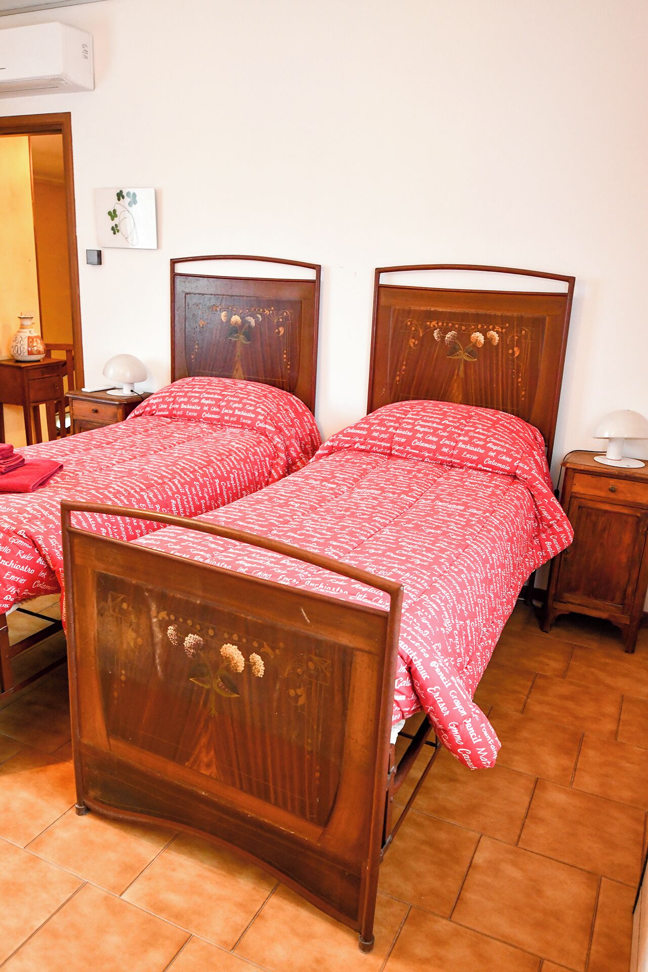 Dos camas de madera con ropa de cama estampada en rojo, una mesita de noche con lámpara y suelo de baldosas.