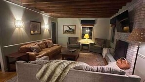 Ein gemütliches Wohnzimmer mit einer Couch, Sesseln und einem Couchtisch. Ein Kamin und eine Lampe auf einem Beistelltisch sind ebenfalls vorhanden.