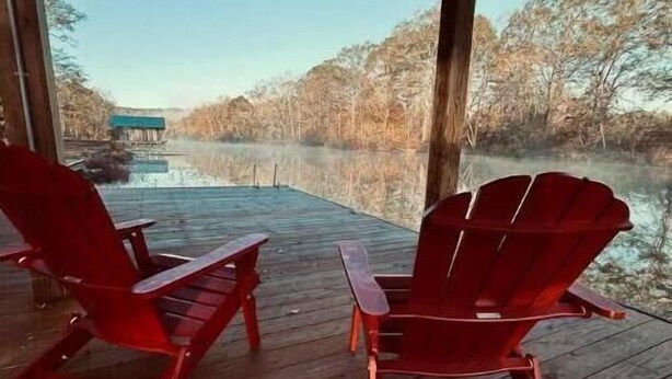 Eine hölzerne Veranda mit zwei roten Adirondack-Stühlen, die auf einen ruhigen See blicken, umgeben von Bäumen.