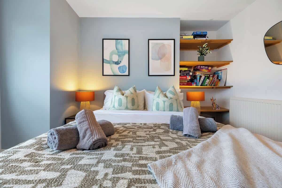 A bedroom with a bed, pillows, a bedside table, a lamp, a shelf with books, and two framed pictures on the wall.