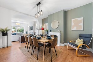 Un comedor con una mesa de madera dispuesta para cuatro personas, rodeada de sillas. Hay una chimenea con un espejo encima y una silla moderna con cojín a la derecha.