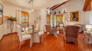 Amplia sala de estar con una mesa de centro de vidrio, sofás blancos, sillas de mimbre y un comedor con mesa y sillas de vidrio.