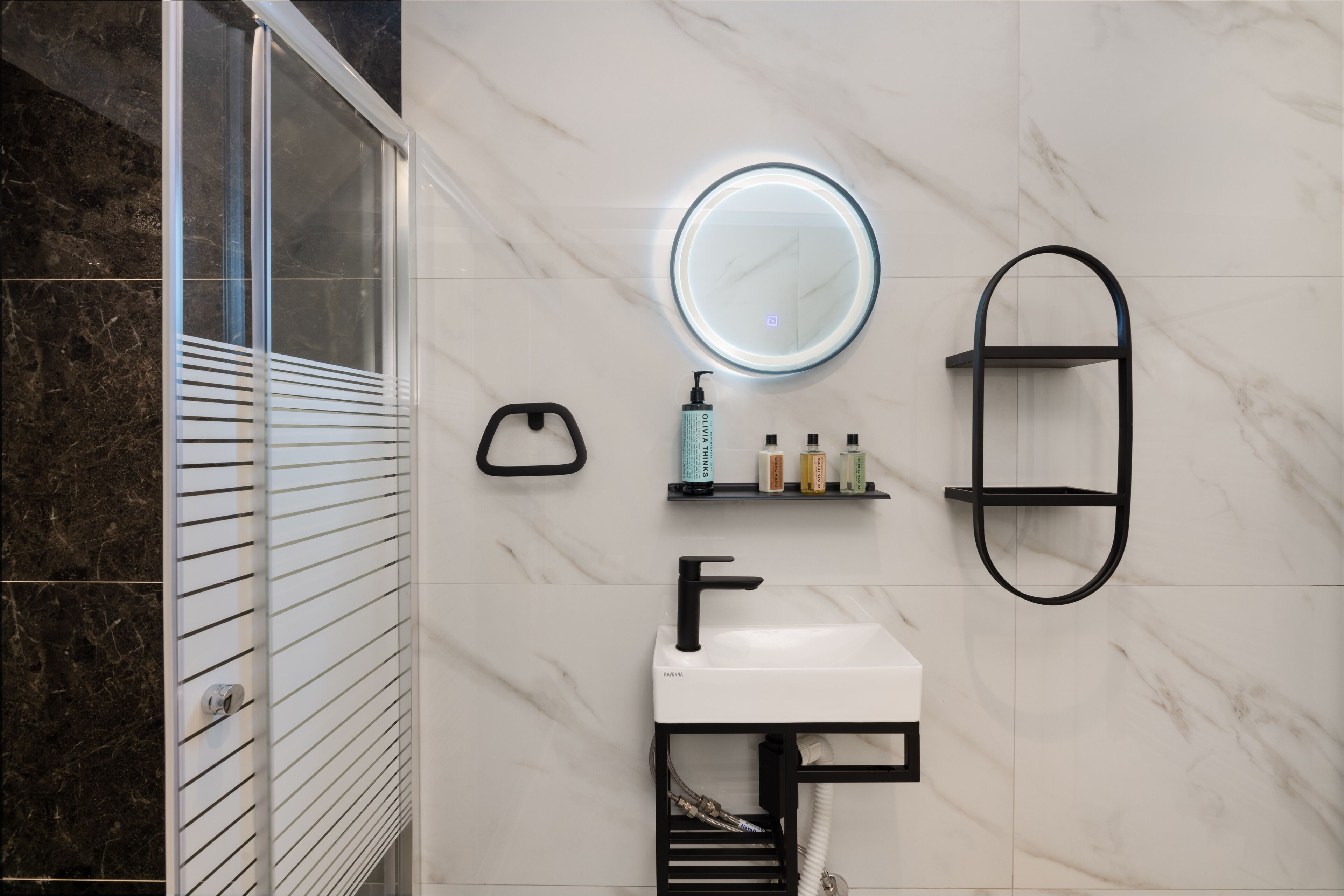 A modern bathroom with a glass shower enclosure, a white sink with a black faucet, a round mirror with a light, and a black towel rack.