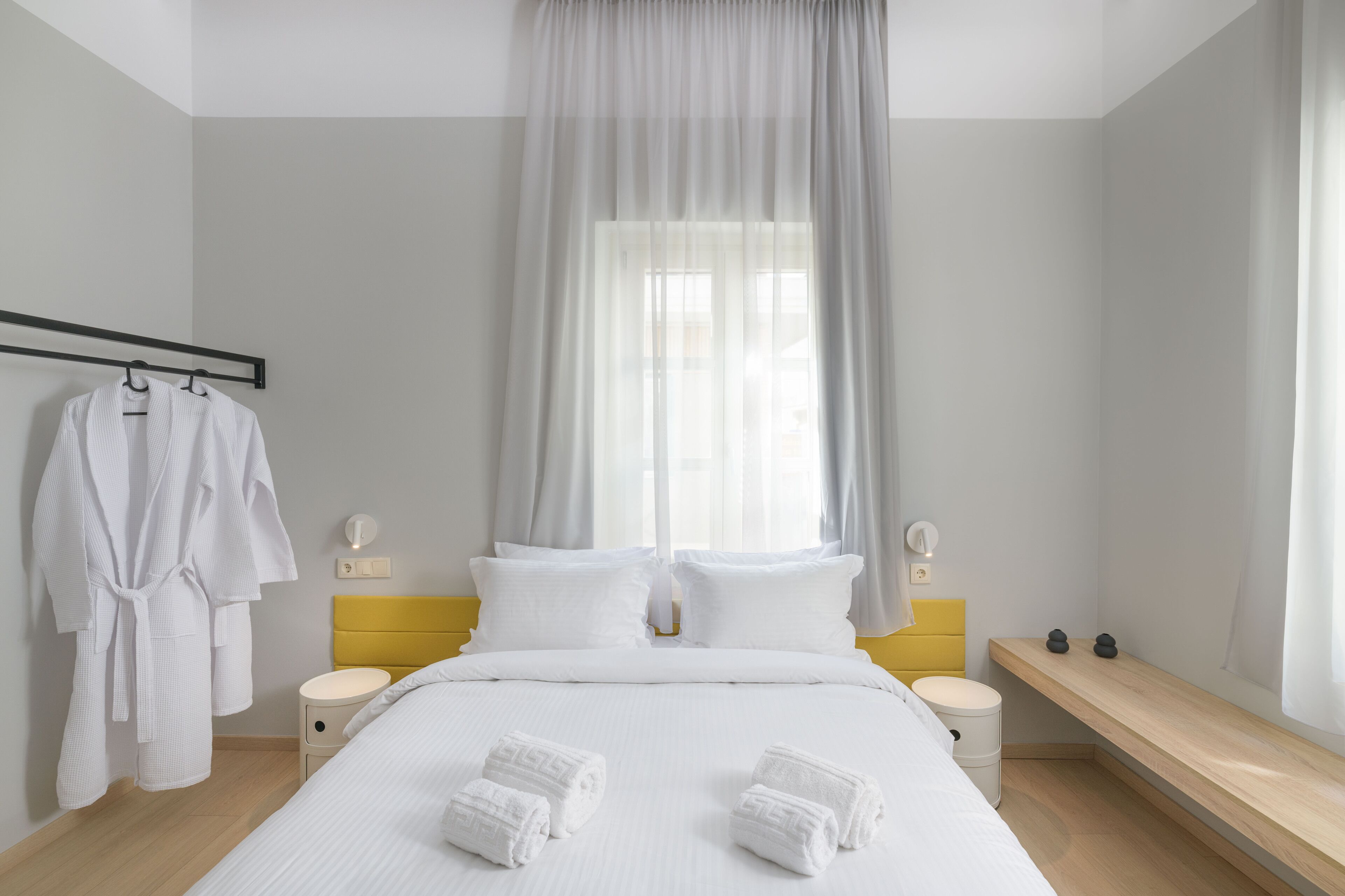 Un dormitorio moderno con una cama, dos mesitas de noche, un banco y una ventana con cortinas traslúcidas.