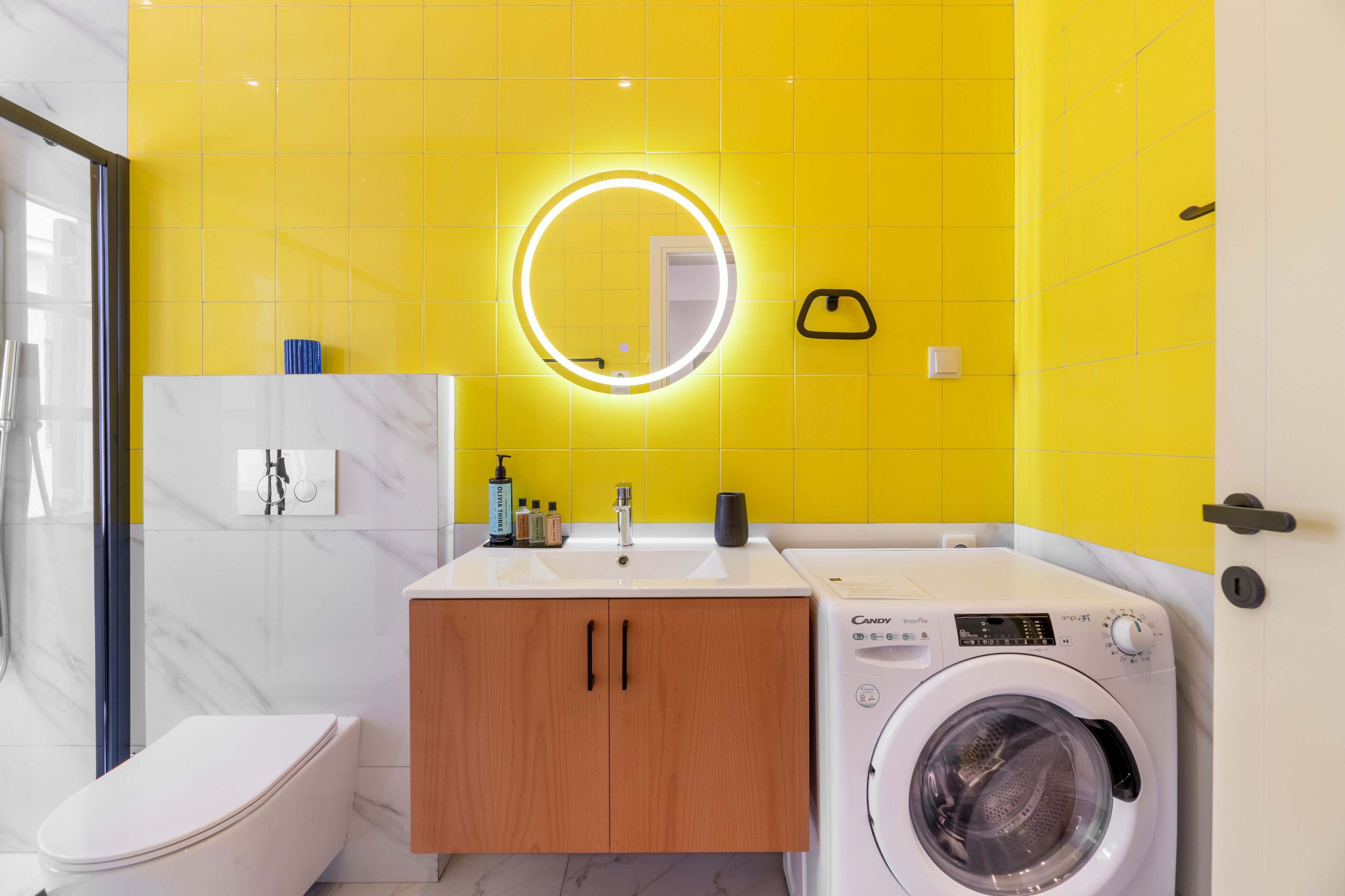 A modern bathroom with a yellow tiled wall, a round mirror with underglow, a white sink, wooden cabinets, a white toilet, and a front-loading washing machine.