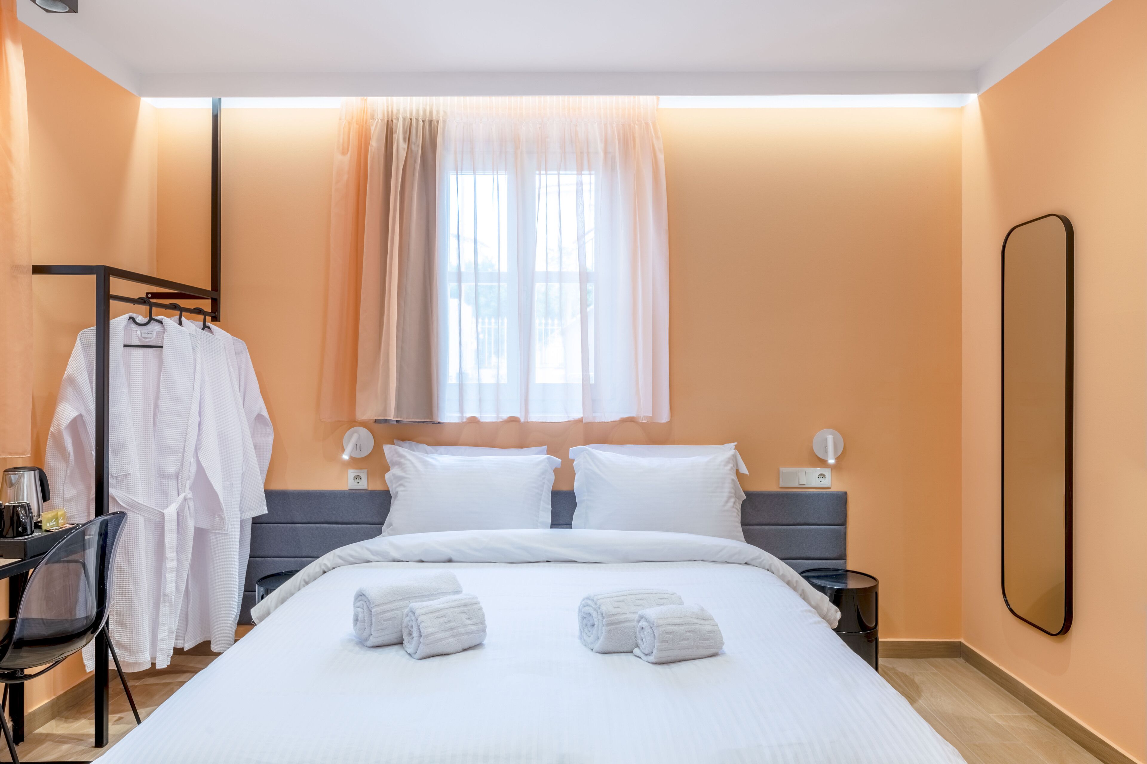 A hotel room with a bed, two bedside tables, a desk with a chair, a mirror, and a window with curtains.