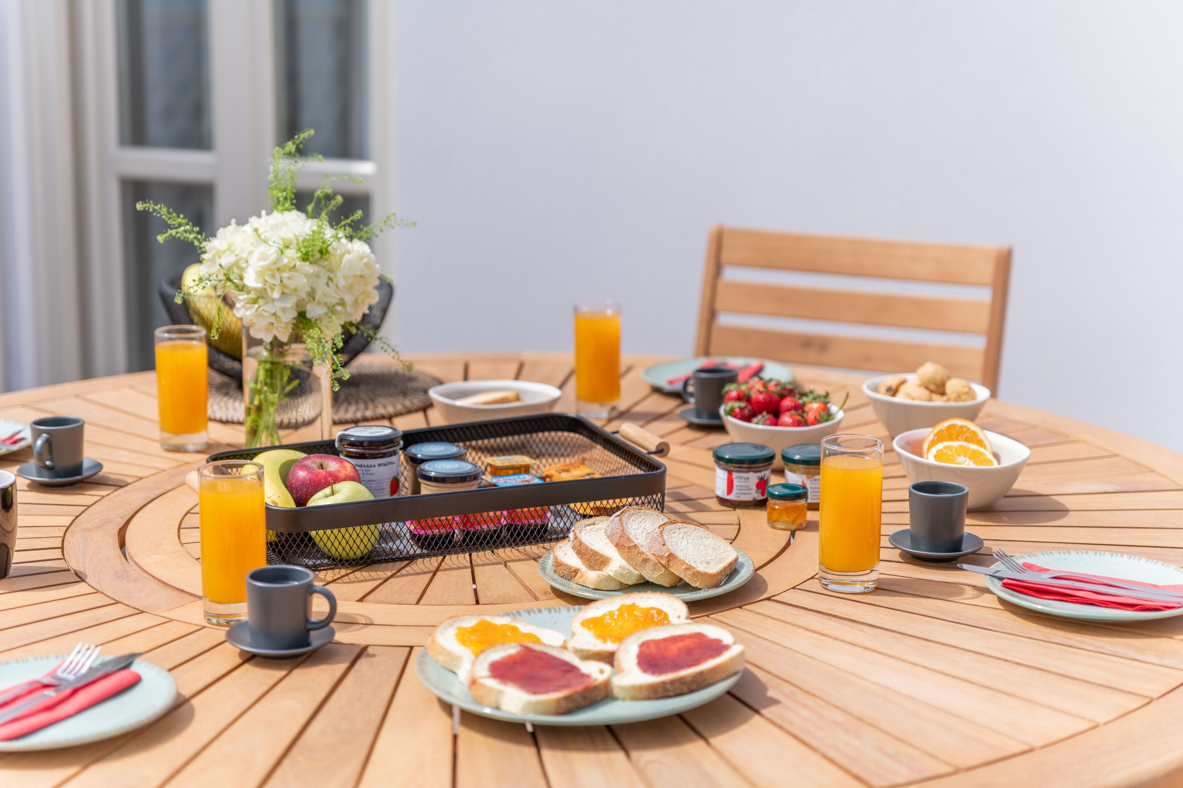Una mesa redonda de madera dispuesta con alimentos para el desayuno, como tostadas, fruta y jugo, con un florero en el centro.