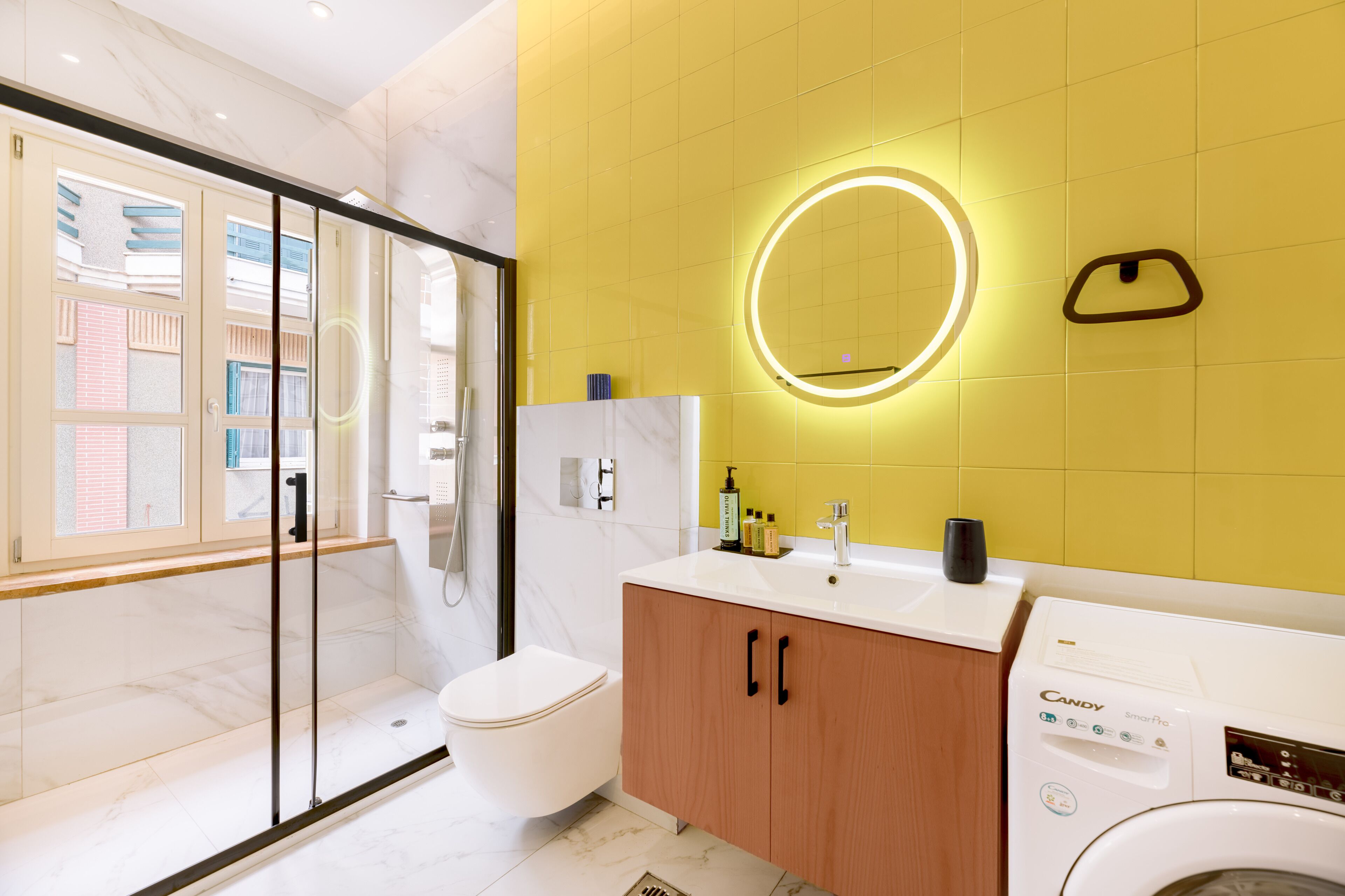 A modern bathroom with a glass shower enclosure, a toilet, a vanity with a mirror, and a Candy SmartPro washing machine.