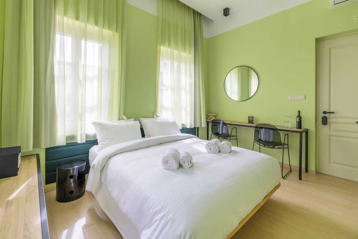 A hotel room with a bed, a desk, a chair, a trash bin, and a door.