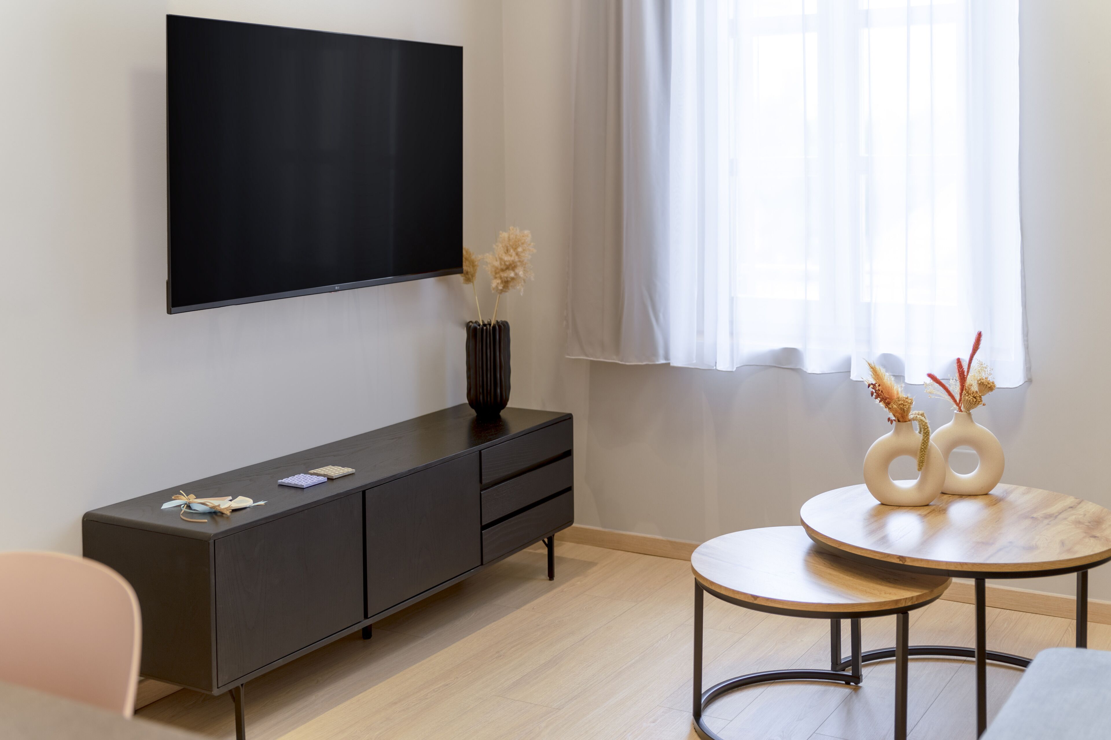 A modern living room with a flat-screen TV mounted on the wall, a dark wooden cabinet with drawers, a round coffee table with two white vases, and a light-colored chair.