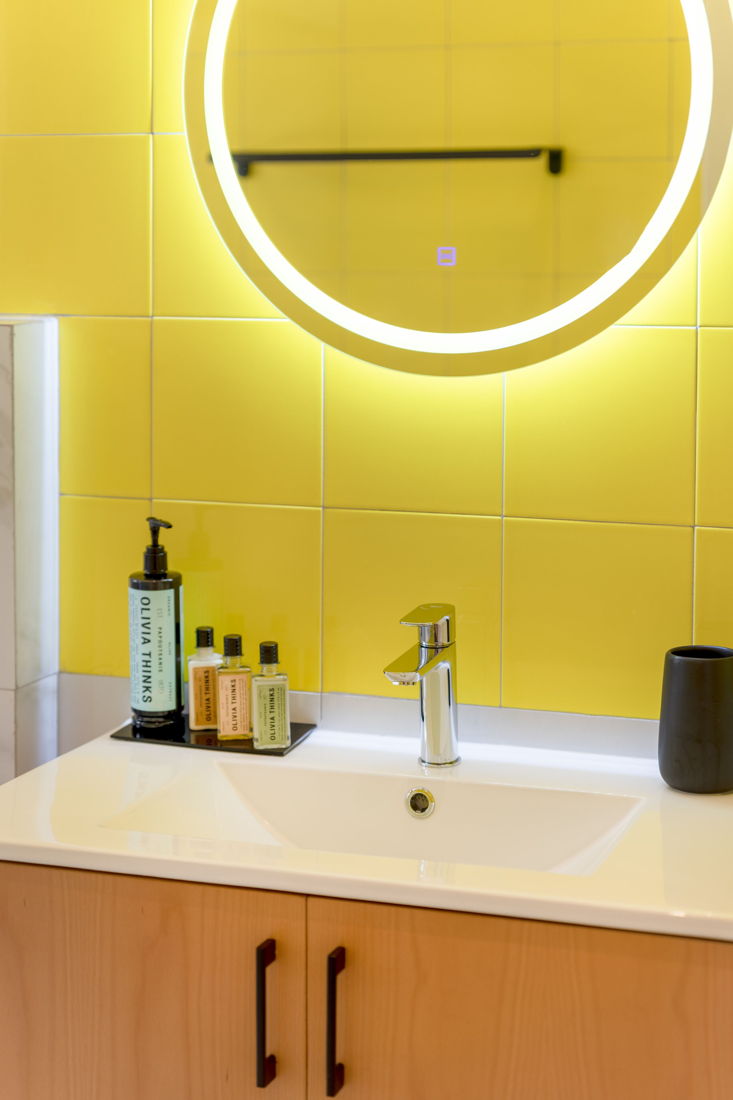 Un baño con un lavamanos blanco, un espejo con luz circular y artículos de aseo en la encimera.