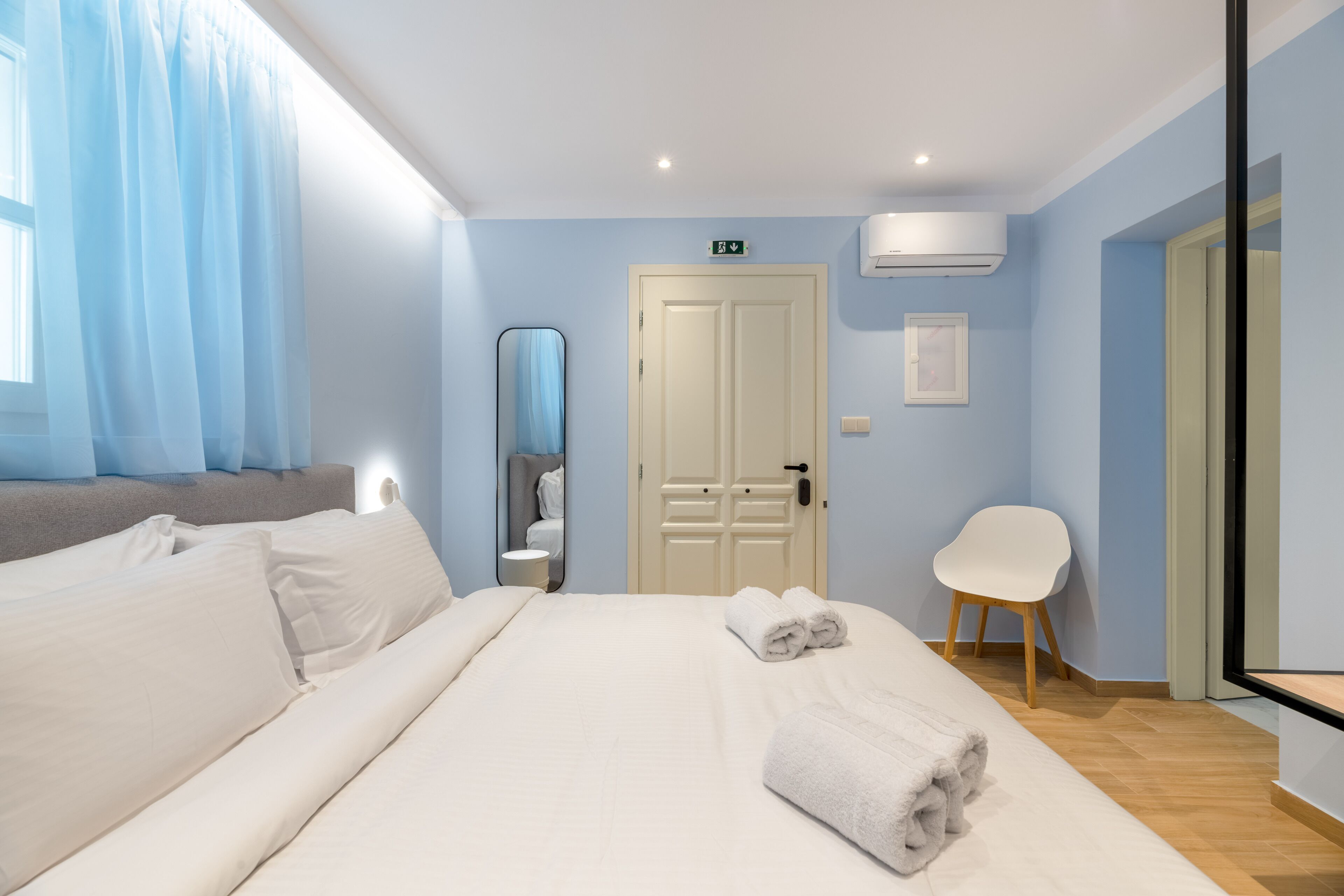 A hotel room with a bed, a chair, a mirror, and a door with an exit sign.