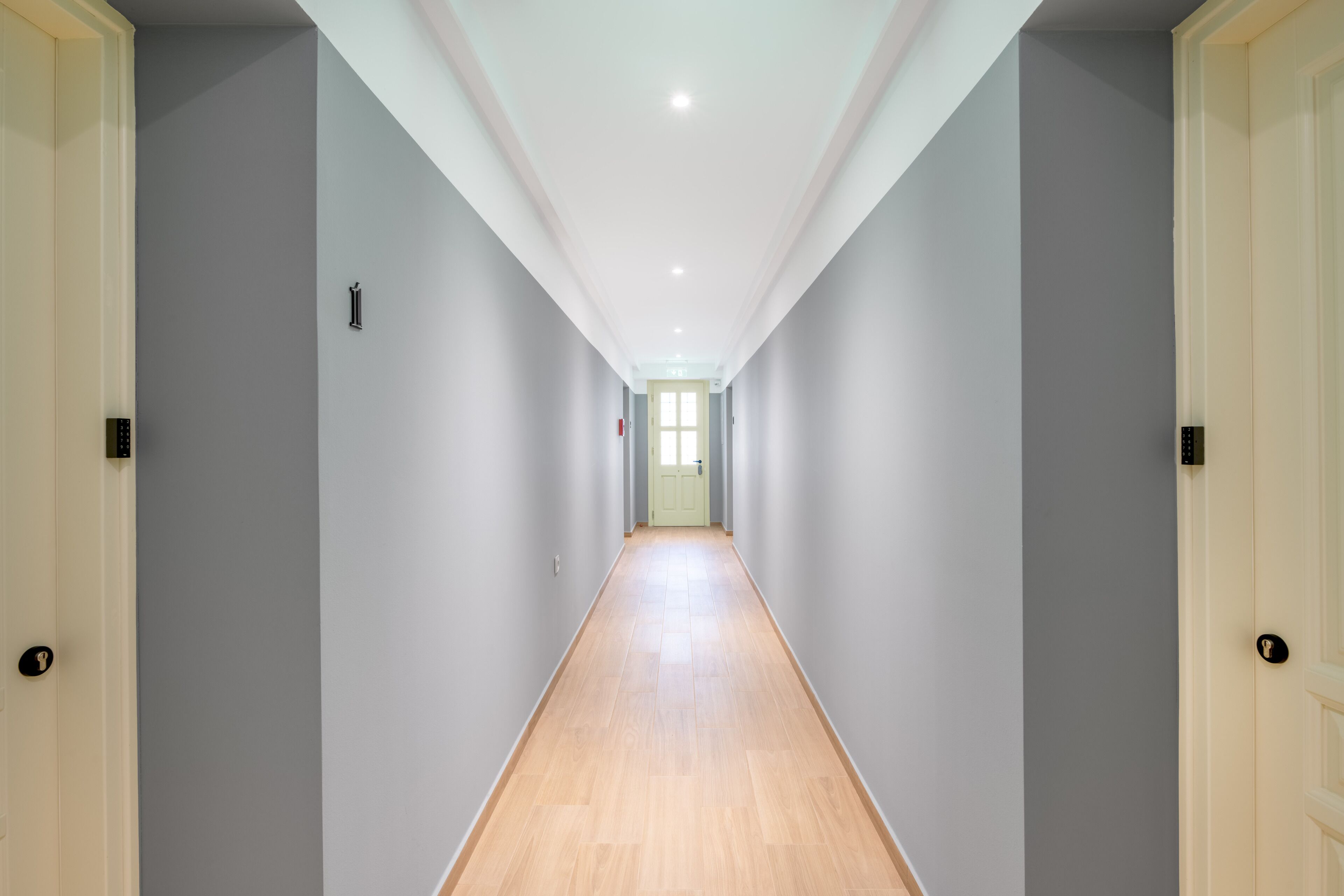 Un pasillo largo y estrecho con paredes gris claro y suelo de madera, que conduce a una puerta al final.