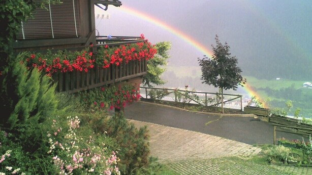 A building with a balcony adorned with red flowers, a paved pathway, and a view of a rainbow over a valley.