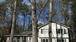 A white two-story house with a brick foundation, surrounded by tall trees. - Pet Friendly Suburban Retreat: Modern Comforts for Families w/ firepit (Marietta)