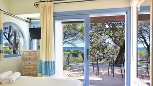 A bedroom with a bed, a wooden dresser, a TV mounted on the wall, and a view of the outdoors through a glass door. - Villaggio Colostrai (Muravera)