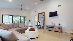 Ein geräumiges Wohnzimmer mit einem beigen Sofa, einem weißen Hocker, einem Holz-TV-Möbel und einem großen Spiegel.