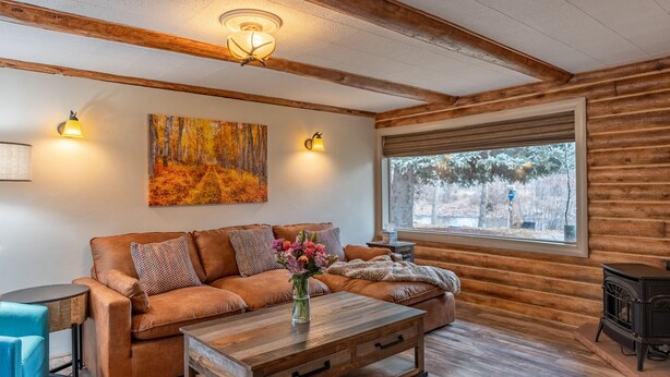 Ein gemütliches Wohnzimmer mit einem braunen Ledersofa, einem Holztisch mit Blumen in einer Vase, einem Bild an der Wand und einem großen Fenster mit Blick auf Bäume.
