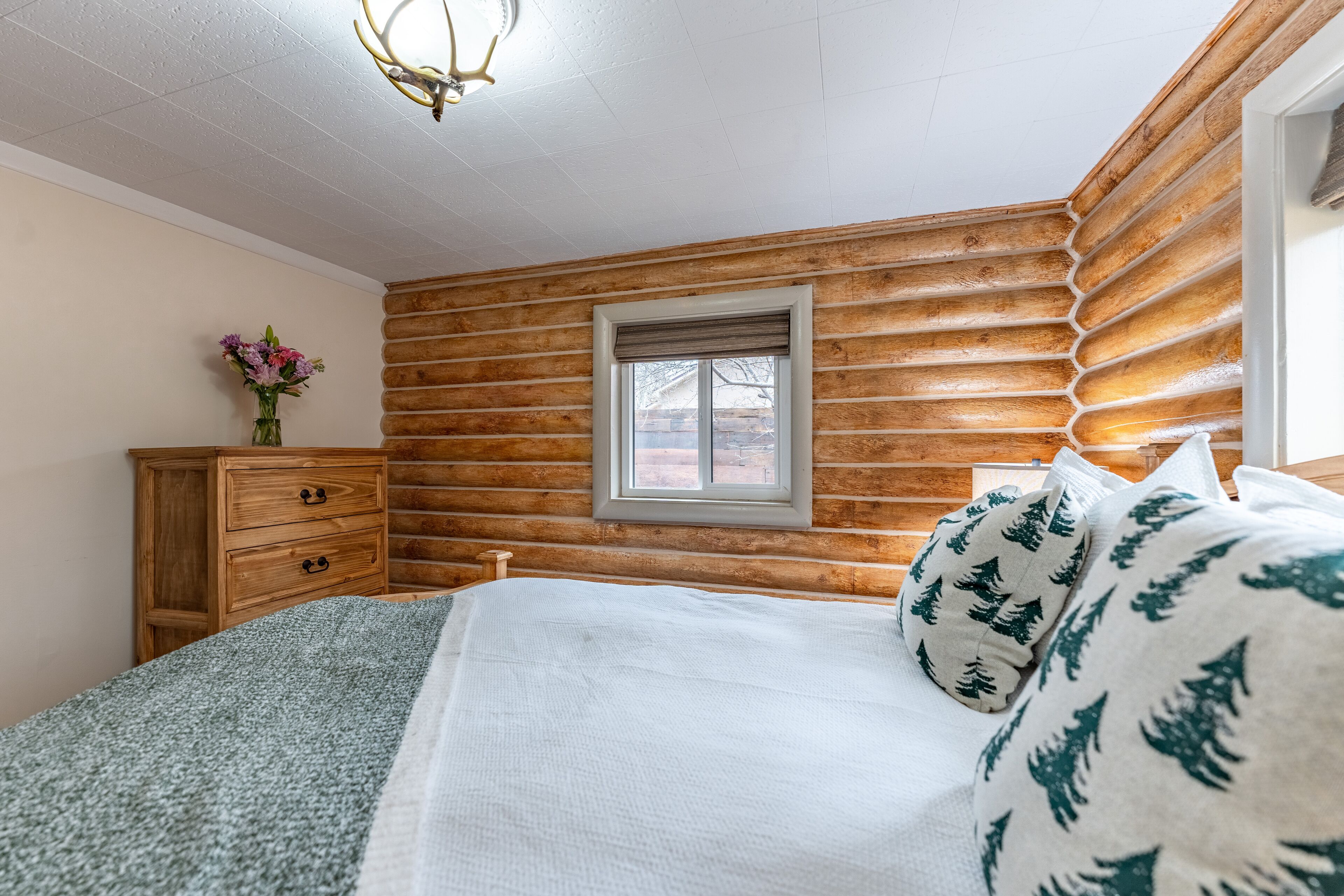 Een slaapkamer met een wand van houten boomstammen, een raam met uitzicht, een houten dressoir en een bed met een witte deken en kussens.