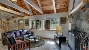 A cozy living room with a stone fireplace, wooden ceiling beams, a leather sofa, and large windows offering a view of the forest.