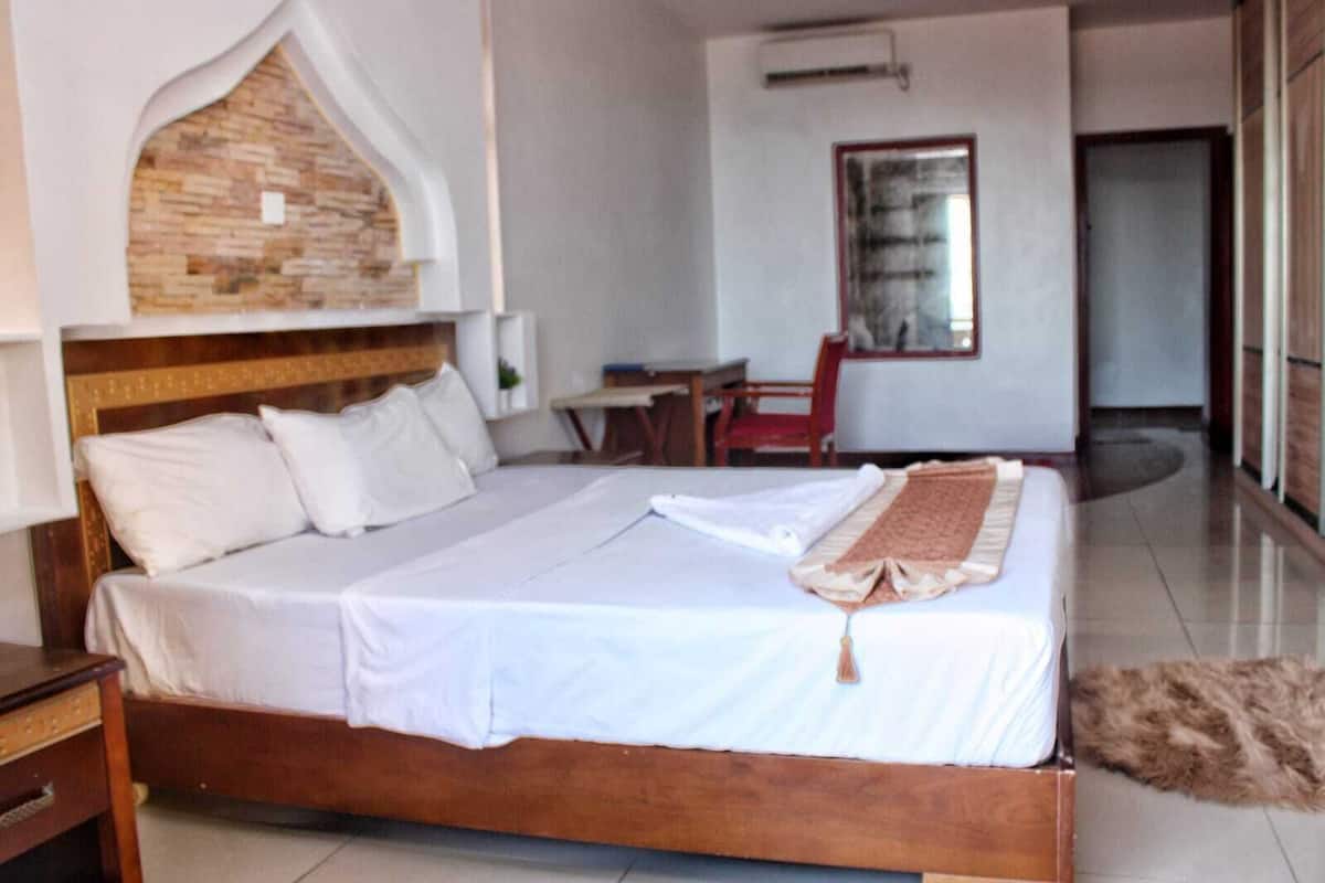 A hotel room with a large bed, a wooden headboard, a small table, a chair, and a view of the outside.