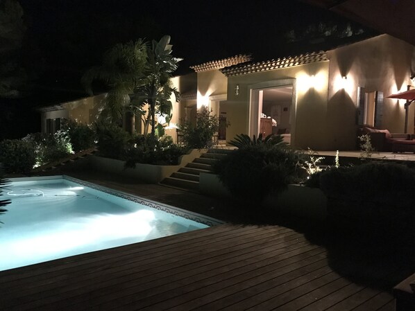 Ein Nachtblick auf einen Poolbereich mit Holzterrasse, umgeben von üppiger Vegetation und einem Gebäude mit beleuchteter Fassade.