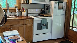 A compact kitchen with a microwave, oven, and refrigerator, wooden cabinets, and a window with a view of greenery. - Whimsical Creekside Cabin (Vesuvius)