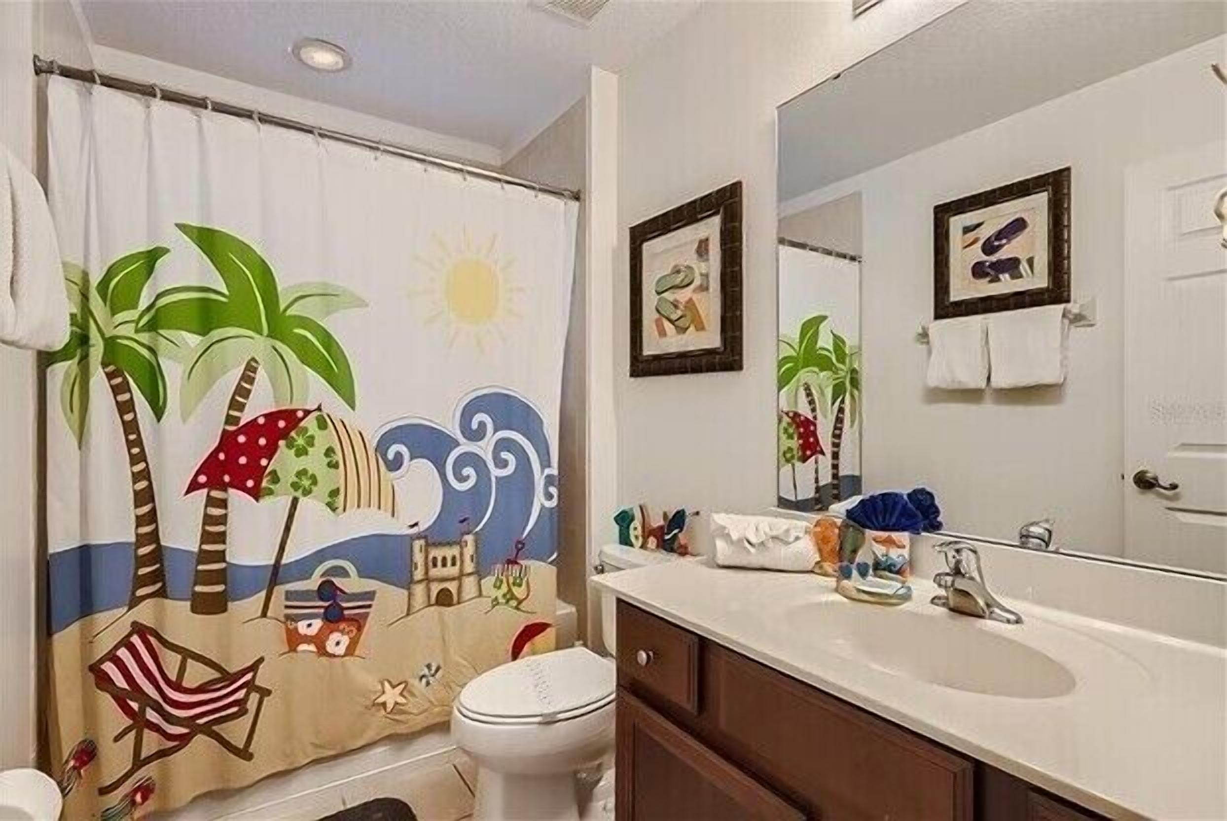 A bathroom with a beach-themed shower curtain, a white toilet, a sink with a mirror, and a cabinet.