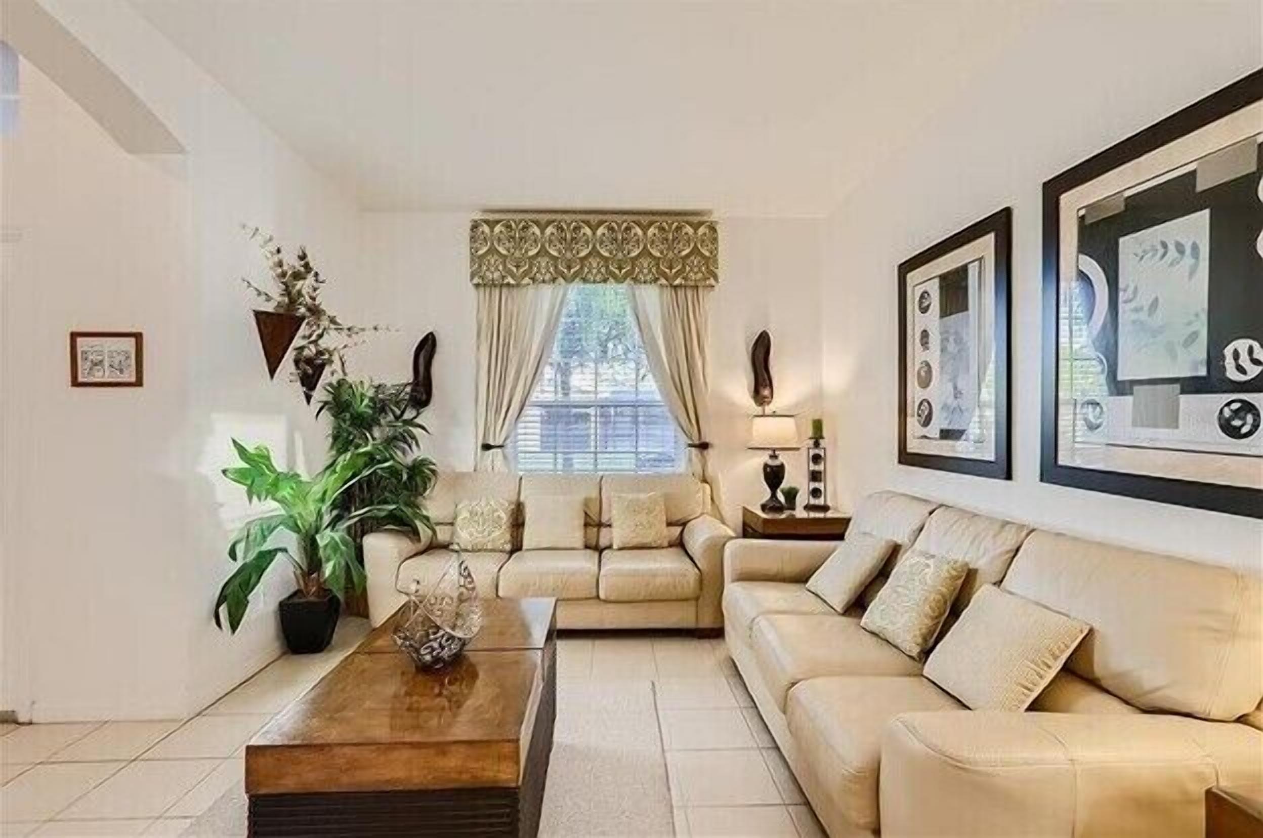 A living room with a beige sofa, a coffee table, and a large window with curtains. There are framed pictures on the wall and a potted plant on the floor.