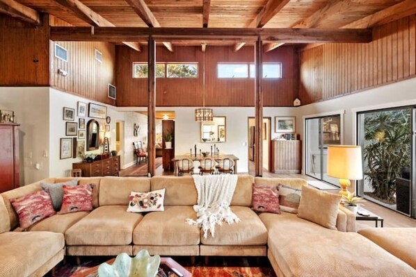 A spacious living room with a large sectional sofa, wooden ceiling beams, and a dining area in the background. - Mid-mod, boho home w/ a pool! (St. Augustine)