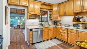 A kitchen with wooden cabinets, a dishwasher, a microwave, and a dining area visible through an open doorway. - Mid-mod, boho home w/ a pool! (St. Augustine)